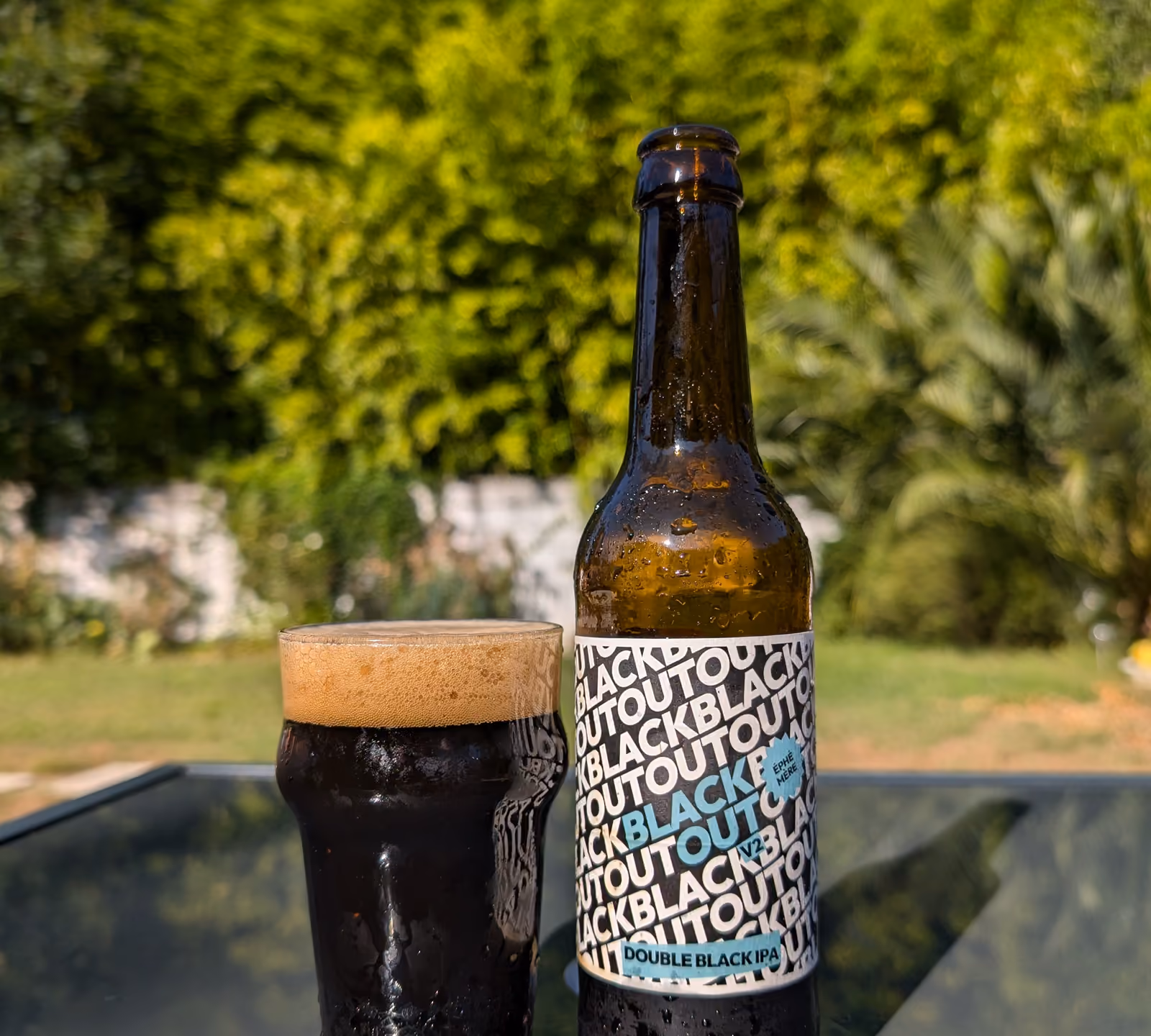 Bouteille de Black Out, double black IPA, et un verre de bière noire posés dehors sur une table.